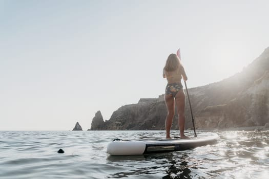 Woman paddleboarding in calm ocean water, facing away from the camera, with rocky cliffs in the background.