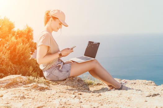 A young woman wearing a cap, sitting on a cliff, works on her laptop with the ocean in the background.