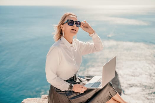 Business woman on nature in white shirt and black skirt. She works with an iPad in the open air with a beautiful view of the sea. The concept of remote work