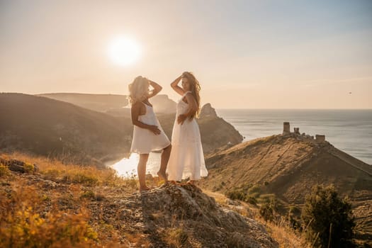 Two young girls are standing on a hillside, one of them wearing a white dress. The sun is shining brightly, creating a warm and inviting atmosphere. The girls seem to be enjoying their time together