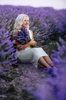 Blonde woman poses in lavender field at sunset. Happy woman in white dress holds lavender bouquet. Aromatherapy concept, lavender oil, photo session in lavender.