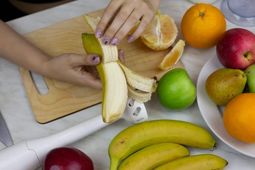 Female hands peeling banana on kitchen table with fruits on background. Girl preparing sports fruit cocktail. Healthy nutrition and lifestyle at home