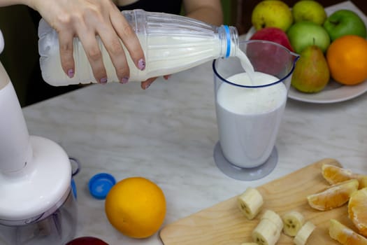 Female hands pouring bottle of milk into measuring cup for blending, making sports drink from fruits and milk with vitamins, healthy homemade milkshake with fresh fruits