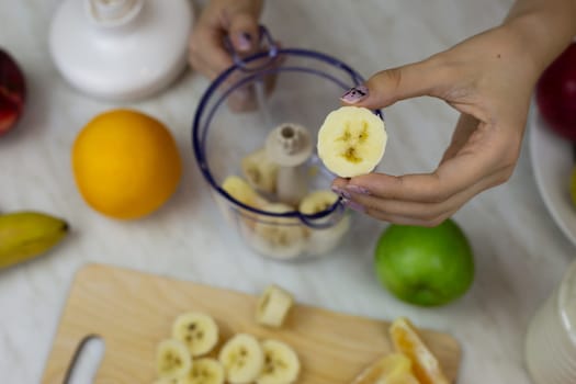 Piece of banana in hand of sporty girl against background of blender with fruits while preparing a sports cocktail. Healthy drink with vitamins from fruits and milk in blender at home kitchen