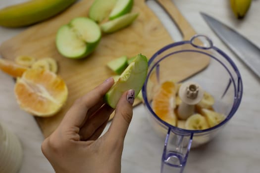 Girl holding piece of green apple in hand against background of blender for chopping fruits while preparing sports healthy cocktail using simple recipe at home
