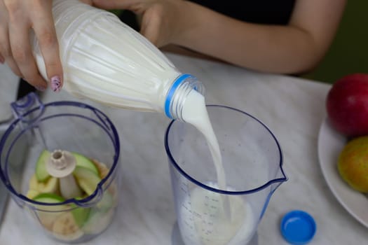 Girl hands pour milk from bottle into measuring cup to add to fruit in blender. Recipe for simple healthy fruit milkshake at home, ingredients for vitamin cocktail