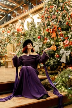 A woman in a purple dress is sitting on a wooden staircase in front of a Christmas tree. She is wearing a fur hat and a necklace. The scene is festive and warm