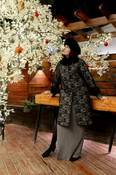 A woman in a black coat stands in front of a Christmas tree. She is wearing a hat and a scarf
