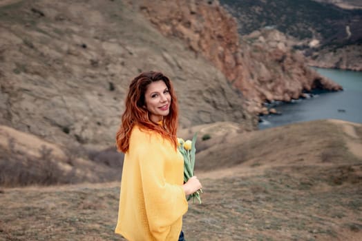 Portrait of a happy woman with long hair against a background of mountains and sea. Holding a bouquet of yellow tulips in her hands, wearing a yellow sweater.