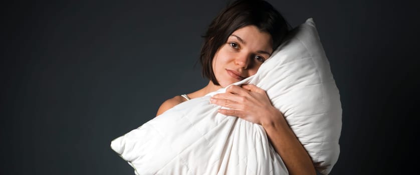 A young girl stands and hugs a white pillow on a gray background, a woman wants to sleep, take care of her health, the quality of sleep.