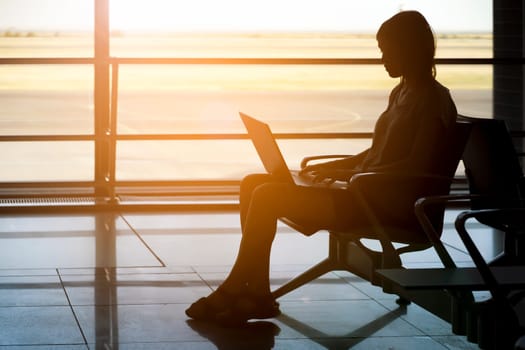 A young girl works on a laptop at the airport while she waits to board a plane. A woman buys tickets, studies and communicates via the Internet, the airport in the background.