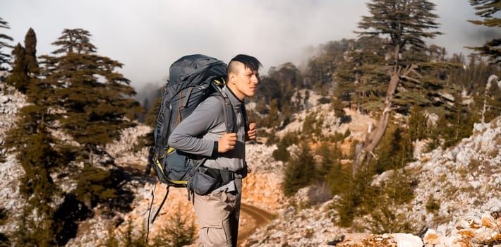 A young man with tourist equipment in a backpack, in trekking pants and a fleece jacket climbs to the top of the mountain. The traveler goes hiking in early spring. Rocks and stones on the background.