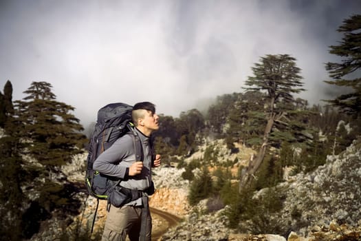 A young man with tourist equipment in a backpack, in trekking pants and a fleece jacket climbs to the top of the mountain. The traveler goes hiking in early spring. Rocks and stones on the background.