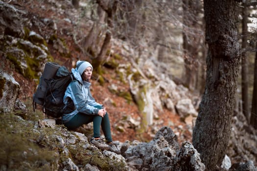 A young girl sits with a backpack and hiking equipment on a stone, rests during her climbing in the mountains and forest, the woman leads an active lifestyle and is involved in mountaineering.