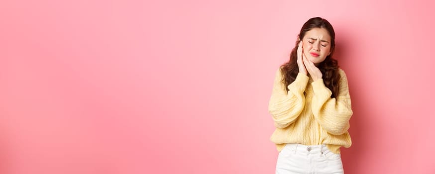 Young woman with terrible toothache, touching swallen cheek and grimacing from painful feeling, having tooth decay, need appointment to dentist, standing against pink background.