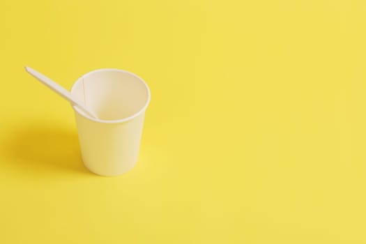 Paper glass with a plastic spoon on a yellow background, top view.