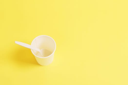 Paper glass with a plastic spoon on a yellow background, top view.