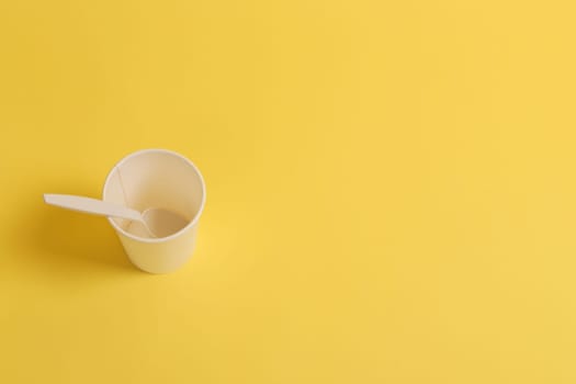 Paper glass with a plastic spoon on a yellow background, top view.