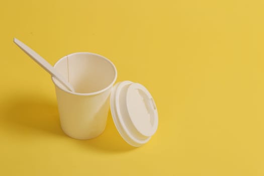 Eco-friendly dishes on a yellow background. Disposable glass with an open lid, with a teaspoon on a yellow background.