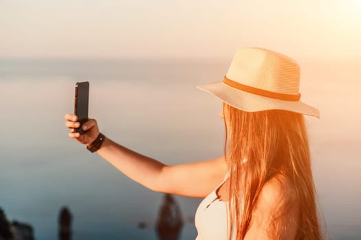 Selfie woman in a hat, white tank top, and shorts captures a selfie shot with her mobile phone against the backdrop of a serene beach and blue sea