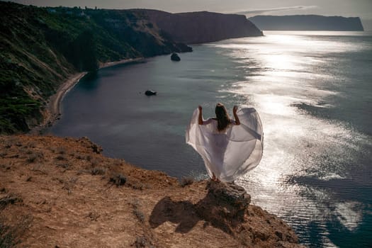 A woman stands on a rocky cliff overlooking the ocean. She is wearing a white dress and her arms are spread wide, as if she is embracing the beauty of the scene. Concept of freedom and serenity