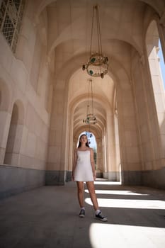 A woman in a white dress walks through a long, arched corridor with a chandelier hanging above.