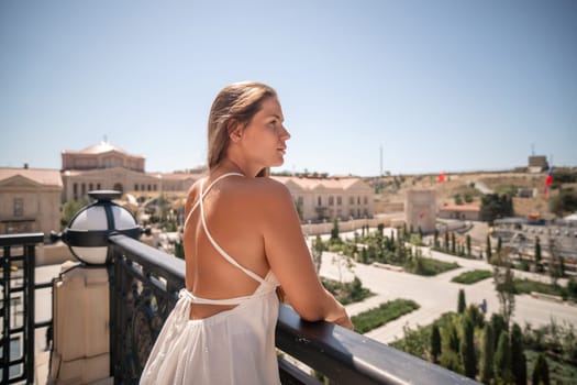 A woman in a white dress stands on a balcony overlooking a city park. She looks out over the cityscape, with her back to the camera. The day is sunny and bright.
