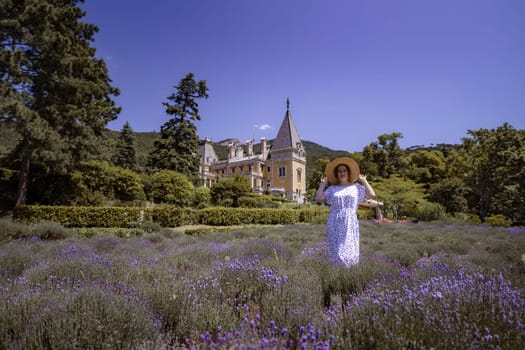 A woman is standing in a field of purple flowers. She is wearing a white dress and a straw hat. The field is surrounded by trees and the sky is clear and blue. The scene is peaceful and serene