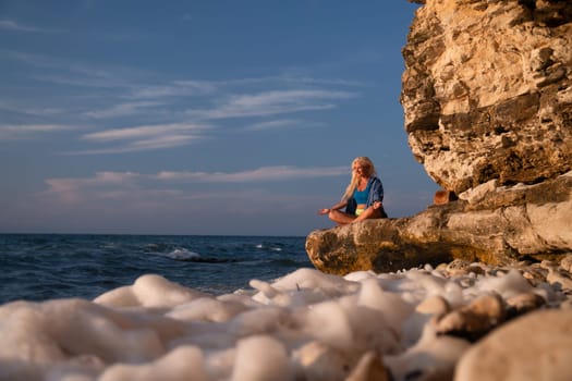 A woman sits cross-legged on a rocky outcrop overlooking the sea, with waves crashing on the shore.