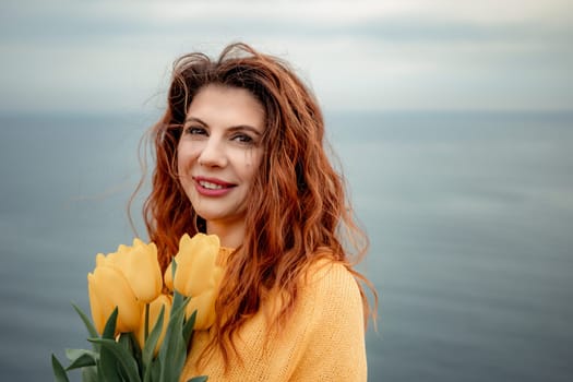 Portrait of a happy woman with hair flying in the wind against the backdrop of mountains and sea. Holding a bouquet of yellow tulips in her hands, wearing a yellow sweater.