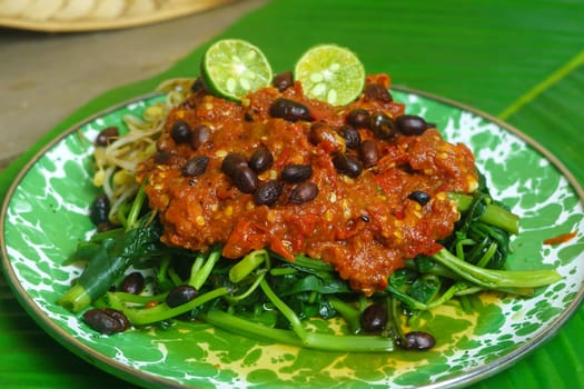 A vibrant plate of Plecing Kangkung, Lombok, Indonesian traditional food. Featuring stir fried water spinach topped with a spicy sambal made from chili peppers, tomatoes, shallots, and garlic, garnished with bean sprouts and lime wedges.
