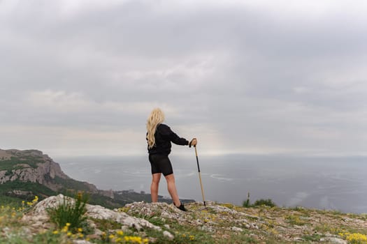 A woman stands on a rocky hill overlooking the ocean. She is holding a walking stick and wearing shorts. The sky is cloudy, and the ocean is visible in the distance