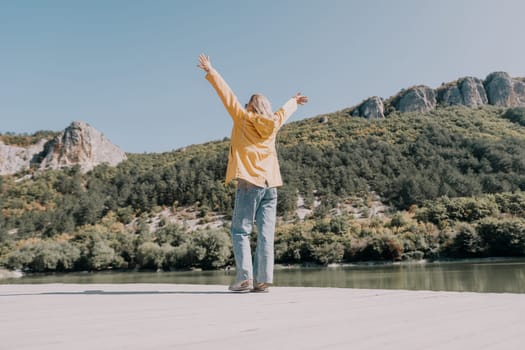 A young woman in a yellow jacket stands on a wooden dock, arms outstretched towards the mountains and lake. She is wearing blue jeans and sneakers, facing a beautiful natural scenery. The sunny, calm atmosphere conveys a sense of freedom and joy.