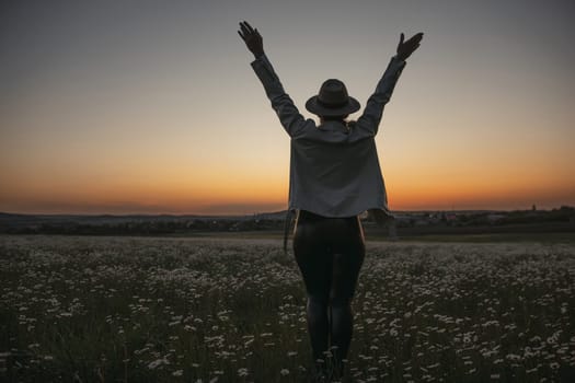 A woman stands in a field of flowers, wearing a hat and a white shirt. She is looking up at the sky, possibly in a state of joy or wonder. The scene is peaceful and serene