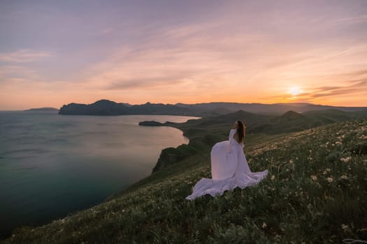 A woman in a white dress stands on a hill overlooking a body of water. The sky is a mix of orange and blue, creating a serene and peaceful atmosphere. The woman is enjoying the view