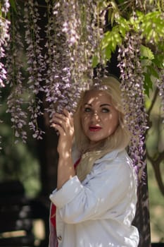 A blonde woman is standing in front of purple flowers. She is wearing a white jacket and red shirt