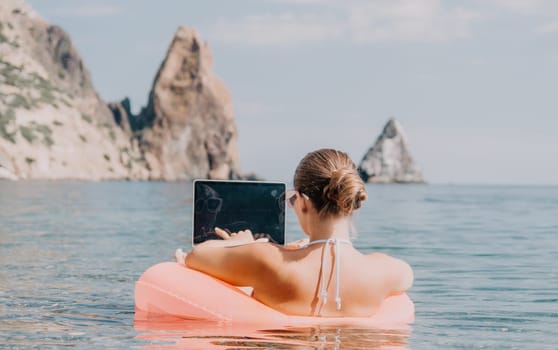 Woman freelancer works on laptop swimming in sea on pink inflatable ring. Pretty lady typing on computer while floating in the sea on inflatable donut at sunset. Freelance, remote work on vacation