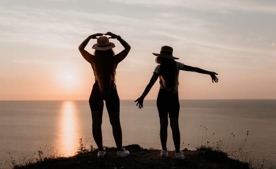 Two women in hats stand on a cliff, silhouetted against a sunset over the ocean.