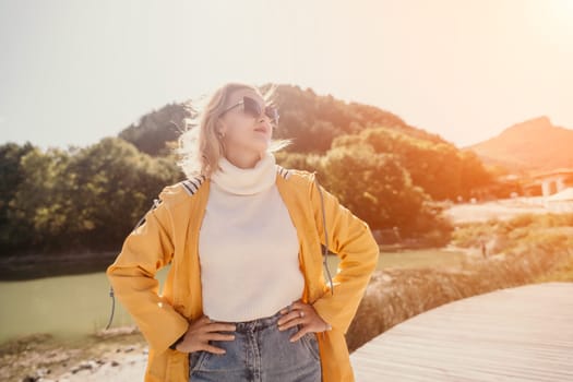 A woman in a yellow coat stands with her hands on her hips, looking up at the sun.
