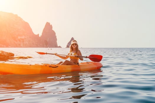 A woman paddles a bright orange kayak in the ocean, with mountains in the distance.