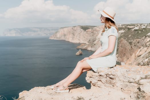 A young woman in a white hat sits on a cliff, looking out at a vast expanse of blue sea.