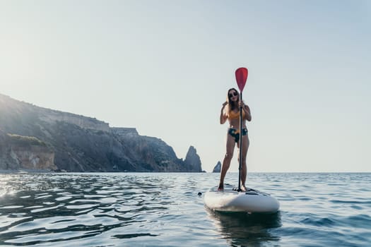 A woman paddles a stand-up paddleboard on calm blue water, with a rocky coastline in the background.