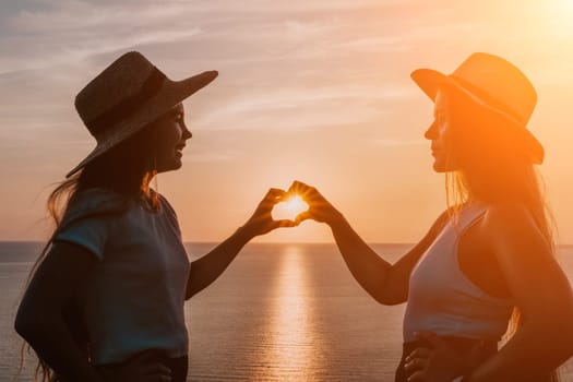 Two women in hats stand in silhouette against the setting sun, creating a heart shape with their hands.