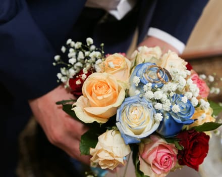 The groom's hands hold the bride's bouquet with red, blue, pink roses, top view.