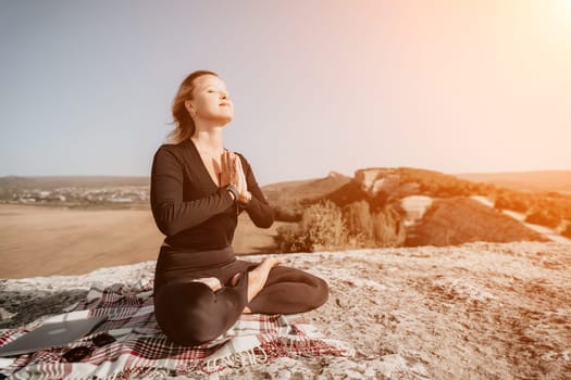 A woman sits in a meditative pose on a mountaintop, with a laptop and blanket nearby. The landscape stretches out behind her, and the sky is clear and blue.