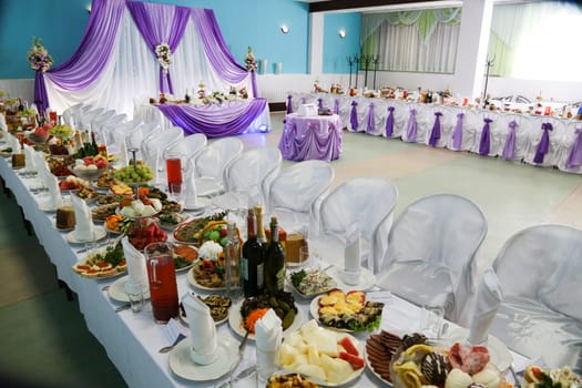 The interior of a large restaurant with wedding decor from curtains, bouquets of flowers, tables with a white tablecloth, a lot of beautiful and varied food on the tables.