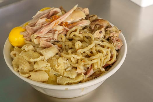 A bowl of Mie Ayam with various toppings, including shredded chicken, meatballs, egg, and vegetables. Served with a separate bowl of soup and condiments.