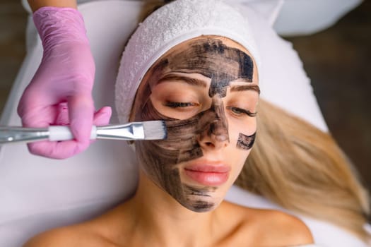 cosmetologist applying black mask on pretty woman face wearing black gloves, gorgeous woman in spa having facial procedures.