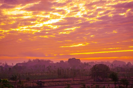 A breathtaking sunrise paints the sky in vibrant hues of orange and pink, casting a warm glow over a rural landscape dotted with trees and fields.