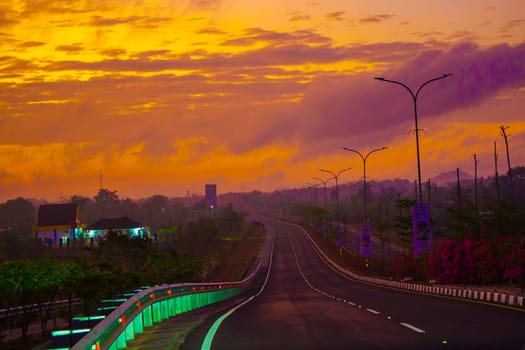 A vibrant sunrise paints the sky above a long, empty road lined with streetlights, leading towards a distant horizon.